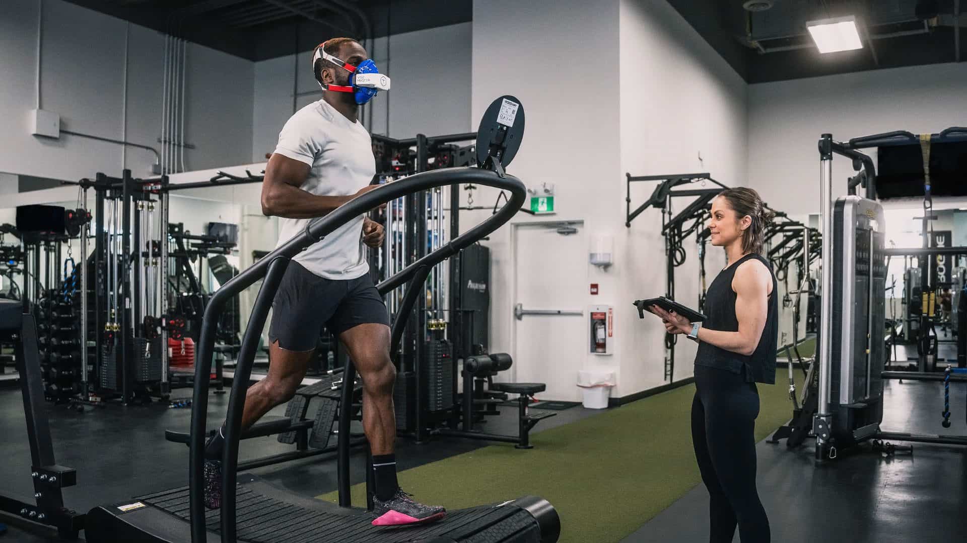 Woman reviewing performance analyzer results on an iPad inside an athletic facility while man runs on treadmill